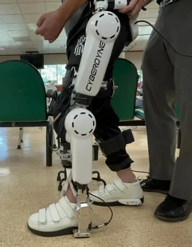 Worker wearing industrial lower-limb powered exoskeleton lifting heavy package in warehouse, with motorized joint actuators at hip and knee joints